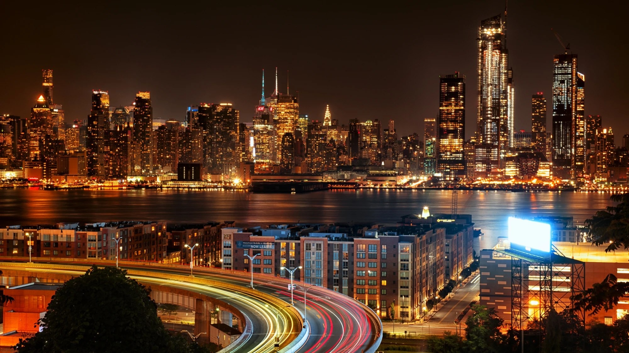 Manhattan skyline at night across the Hudson River with light trails on a curved road